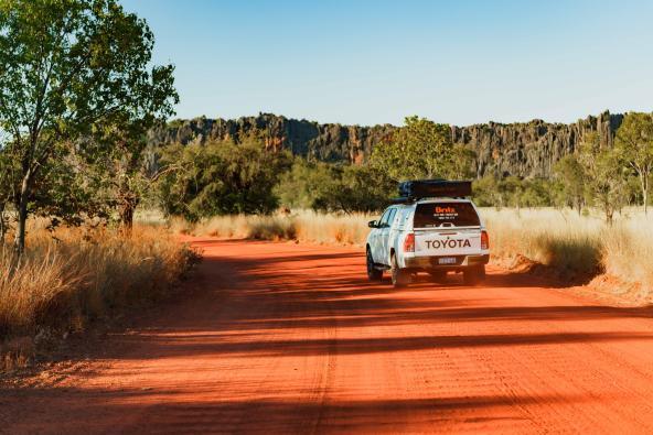 Geländewagen fährt auf roter Schotterstraße in Richtung Windjana Gorge, umgeben von Busch und Felsformationen