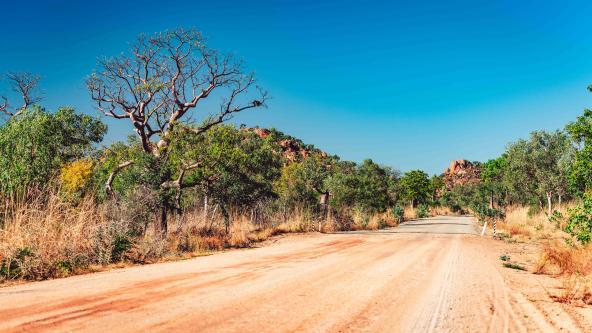 Rote Schotterstraße durch Buschland mit Baobabs und Felsen unter blauem Himmel