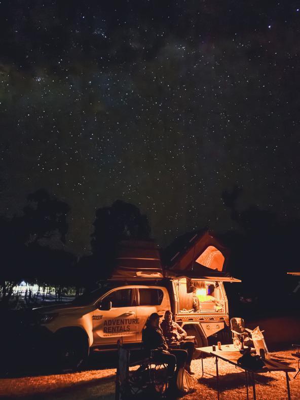 Zwei Personen sitzen abends vor einem beleuchteten Offroad-Camper unter sternenklarem Himmel