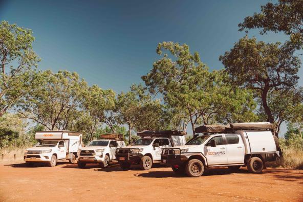 Mehrere Offroad-Camper stehen auf rotem Sandboden im Schatten von Bäumen