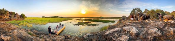 Panoramaaufnahme einer Felsplattform am Wasser mit mehreren Personen, einem kleinen Boot und weitläufigen Feuchtflächen im Sonnenuntergang.