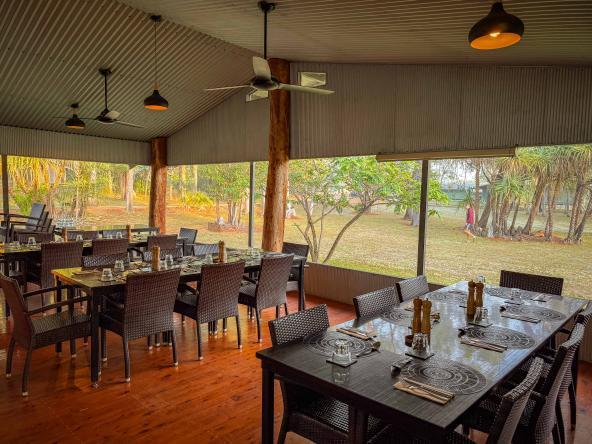 Offener Frühstücksraum mit gedeckten Tischen, Korbstühlen und großem Blick ins umliegende Buschland.