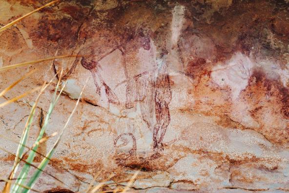 Felsmalerei mit zwei schematischen Figuren und einem kleinen, eingerollten Tiermotiv; einige Grashalme ragen in den Vordergrund.