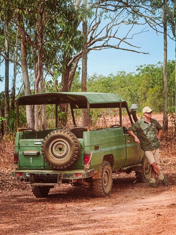 Guide lehnt am offenen grünen Geländewagen am Waldrand; Fahrzeug bereit für den kurzen Transfer zur Landepiste.