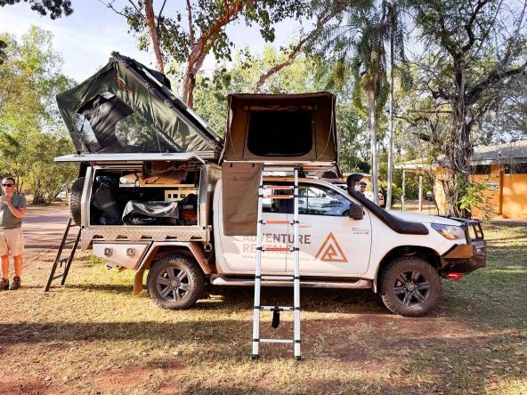 Geländewagen mit zwei geöffneten Dachzelten, Leiter und seitlichen Stauboxen; Personen stehen daneben auf einem Campground.