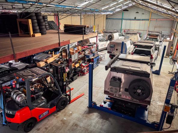 Werkhalle mit mehreren 4WD-Campern auf Hebebühnen, Regale mit Reifen und Werkzeug, Blick von oben.