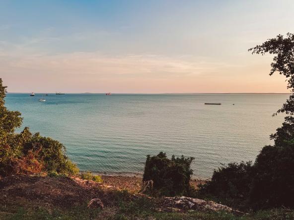Küstenblick im Abendlicht, ruhige See mit mehreren Schiffen am Horizont, Büsche und Felskante im Vordergrund.
