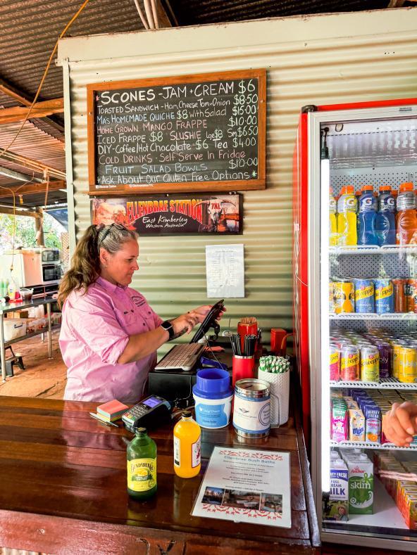 Mitarbeiterin in pinkem Hemd bedient Gäste an der Theke der Ellenbrae Homestead mit Getränkekühlschrank und Menütafel im Hintergrund
