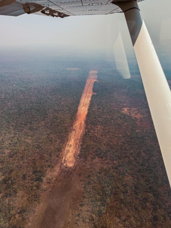 Blick aus einem kleinen Flugzeug auf eine schmale, unbefestigte Landebahn, die sich gerade durch dichtes Buschland zieht.
