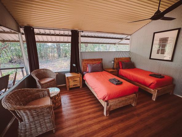 Innenraum mit zwei Einzelbetten, Korbstühlen, Ventilator und großen Fenstern zum Buschland.