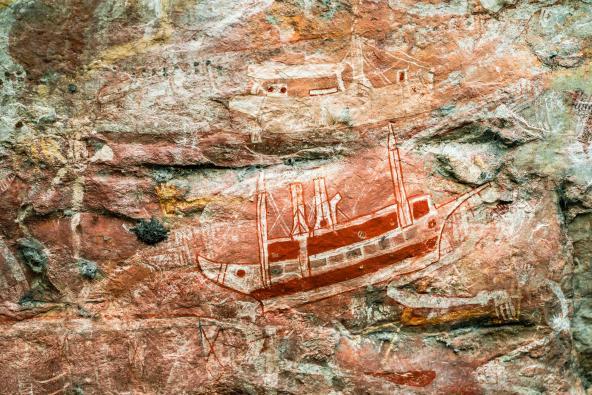 Felsmalerei zeigt ein rot-weiß gemaltes Schiff mit Masten und Aufbauten auf einer unebenen Felswand.
